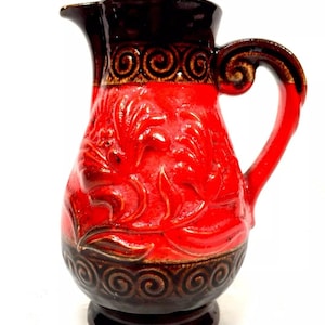 May include: A red and brown ceramic pitcher with a floral design. The pitcher has a wide base and a handle.