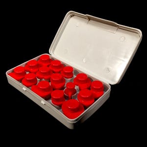 Könnte beinhalten: Eine weiße Kunststoffbox mit einem Klappdeckel, die geöffnet ist, um eine Auswahl an leuchtend roten, abgerundeten Kunststoffteilen zu zeigen. Die Teile variieren in Größe und Form, was auf eine Reihe von Komponenten für einen größeren Artikel oder ein größeres Projekt hindeutet. Der Hintergrund ist schwarz.