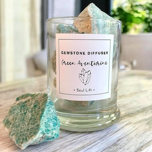 Op de afbeelding: Een glazen edelsteenverspreider gevuld met groene aventurijnstenen. Op het glas staat "Gemstone Diffuser" en "Green Aventurine" met een kristalillustratie. Een losse groene aventurijnsteen ligt ervoor.