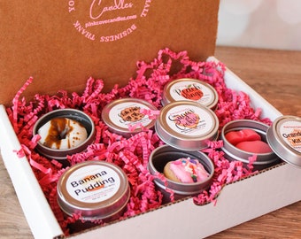 Könnte beinhalten: Eine weiße Geschenkbox mit rosa Seidenpapier im Inneren. Die Box enthält acht kleine, silberne Dosen mit verschiedenen Duftkerzen. Die Kerzen sind thematisch an verschiedene Desserts angelehnt, wie "Sugar Cookie", "Peach Rings" und "Grandma's Kitchen".