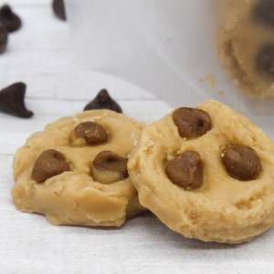 May include: Two round edible cookie dough bites with chocolate chips on a white wooden surface.