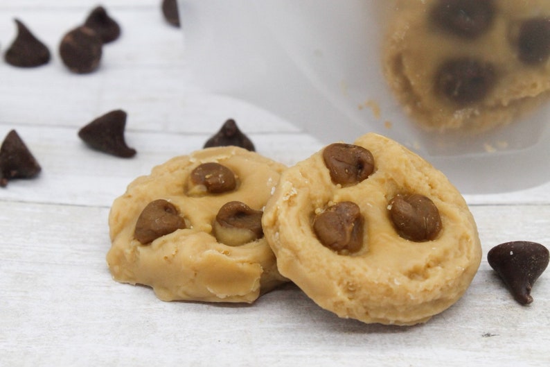 May include: Two round edible cookie dough bites with chocolate chips on a white wooden surface.
