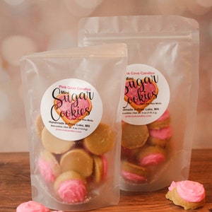 K&ouml;nnte beinhalten: Zwei durchsichtige Plastikt&uuml;ten gef&uuml;llt mit Mini-Zuckerkeks-Duftwachs-Schmelzen. Die Beutel haben Etiketten mit der Aufschrift "Pink Cove Candles" und "Mini Sugar Cookies". Die Wachsschmelzen sind in Form von Zuckerkeksen mit rosa Zuckerguss.