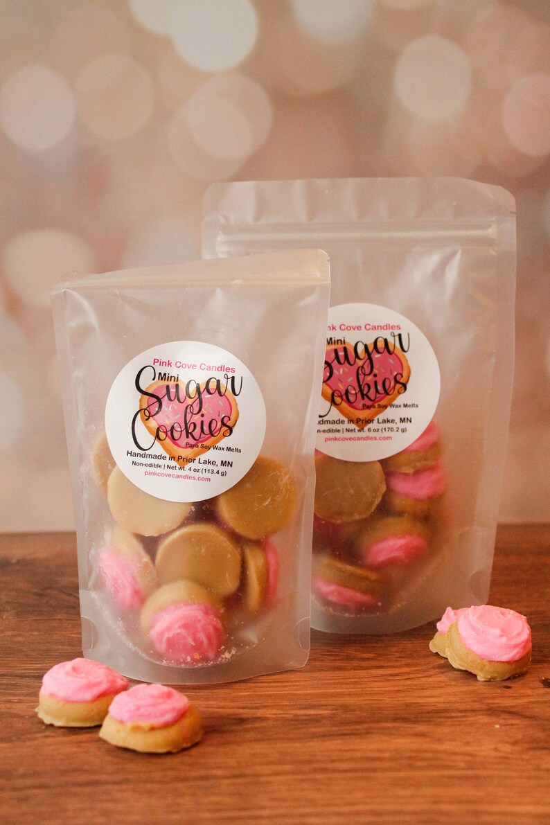 K&ouml;nnte beinhalten: Zwei durchsichtige Plastikt&uuml;ten gef&uuml;llt mit Mini-Zuckerkeks-Duftwachs-Schmelzen. Die Beutel haben Etiketten mit der Aufschrift "Pink Cove Candles" und "Mini Sugar Cookies". Die Wachsschmelzen sind in Form von Zuckerkeksen mit rosa Zuckerguss.