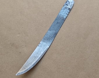 May include: A hand-forged, curved blade knife with a silver-colored blade and a textured, dark gray handle. The knife is angled diagonally across a tan surface. The blade has a sharp edge and a pointed tip.