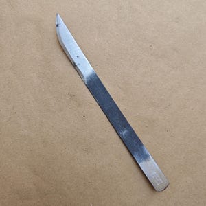 Könnte beinhalten: Ein Metallmeißel mit einer scharfen, abgewinkelten Klinge und einem flachen, rechteckigen Griff. Die Klinge ist silberfarben und der Griff ist dunkelgrau. Der Meißel ist für die Holzbearbeitung oder das Schnitzen konzipiert.