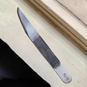 Könnte beinhalten: Ein Metallbrieföffner mit einer langen, flachen Klinge und einem abgerundeten Griff. Die Klinge hat eine leicht gebogene Spitze und ein abgenutztes, gealtertes Aussehen. Der Griff hat ein kleines, gestempeltes Design. Der Brieföffner liegt auf einer hellen Oberfläche.