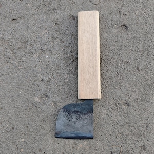 May include: A small, hand-forged knife with a wooden handle. The blade is dark and shows signs of use. The handle is rectangular and made of light-colored wood. The knife is set against a gray, textured surface.