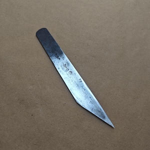 May include: A metal chisel with a rectangular handle and a sharp, angled blade. The blade has a worn, textured appearance with visible markings. The tool is set against a brown background.