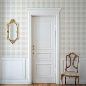 Puede incluir: Una puerta interior blanca con manilla de lat&oacute;n en un marco blanco, flanqueada por frisos blancos. A la izquierda cuelga un espejo ovalado con marco dorado, y a la derecha hay una silla de madera con asiento beige sobre papel pintado a cuadros.