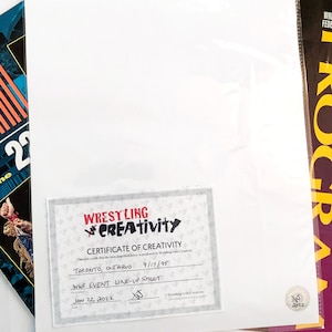 Puede incluir: Un certificado blanco de Wrestling with Creativity, con el texto "WWF EVENT LINE-UP SHEET" y la fecha "22 de enero de 2022". El certificado est&aacute; sobre una hoja blanca, con una revista y un gr&aacute;fico azul y naranja en el fondo.