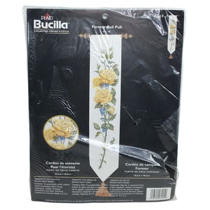 Pode incluir: Um kit de ponto de cruz contado para um marcador de página com rosas amarelas, folhas verdes e flores azuis. O kit inclui tecido Aida de 14 pontos, fio de bordar, dois separadores de fio, uma agulha, instruções e um diagrama. O kit é intitulado "Forever Bell Pull" e é feito nos EUA.
