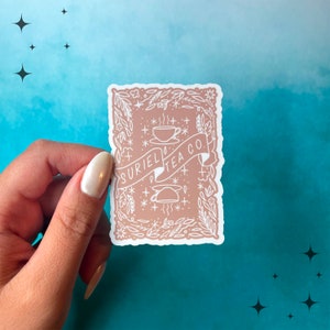 May include: A sticker with a light brown background and white details. The sticker features a teacup, teapot, and the text "SURIEL TEA CO." surrounded by floral and star designs. The sticker is held by a hand against a teal and blue background.