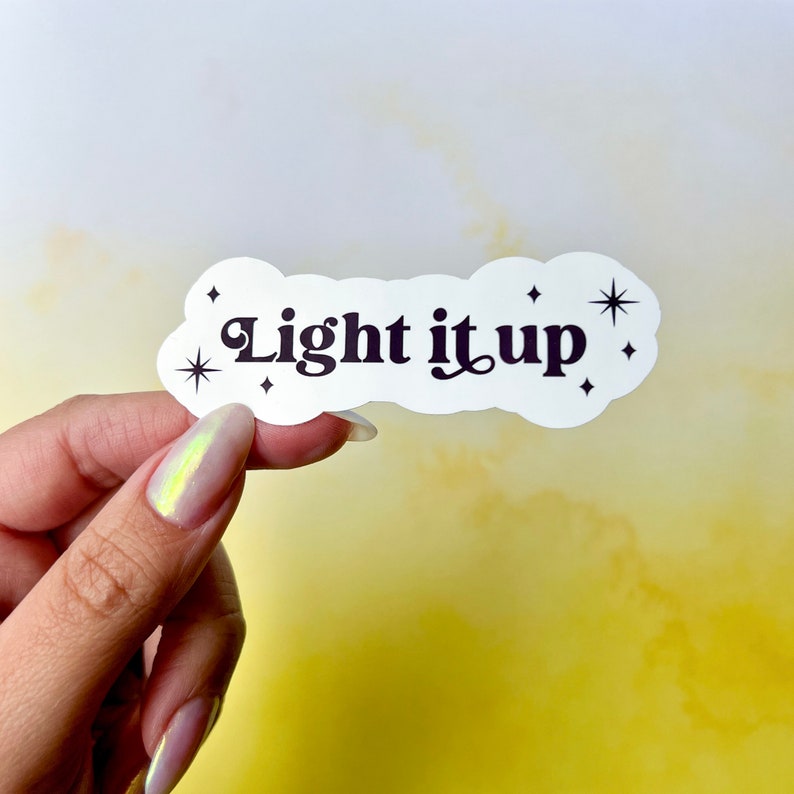 May include: A white sticker in the shape of a cloud with the words "Light it up" in black script. The sticker has small star accents and is held by a hand against a yellow background.