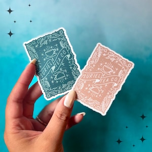 May include: Two rectangular stickers with floral borders and the text "SURIEL TEA CO." are held in a hand. One sticker is teal, the other is a muted peach colour. Both stickers feature a teacup design and star accents.