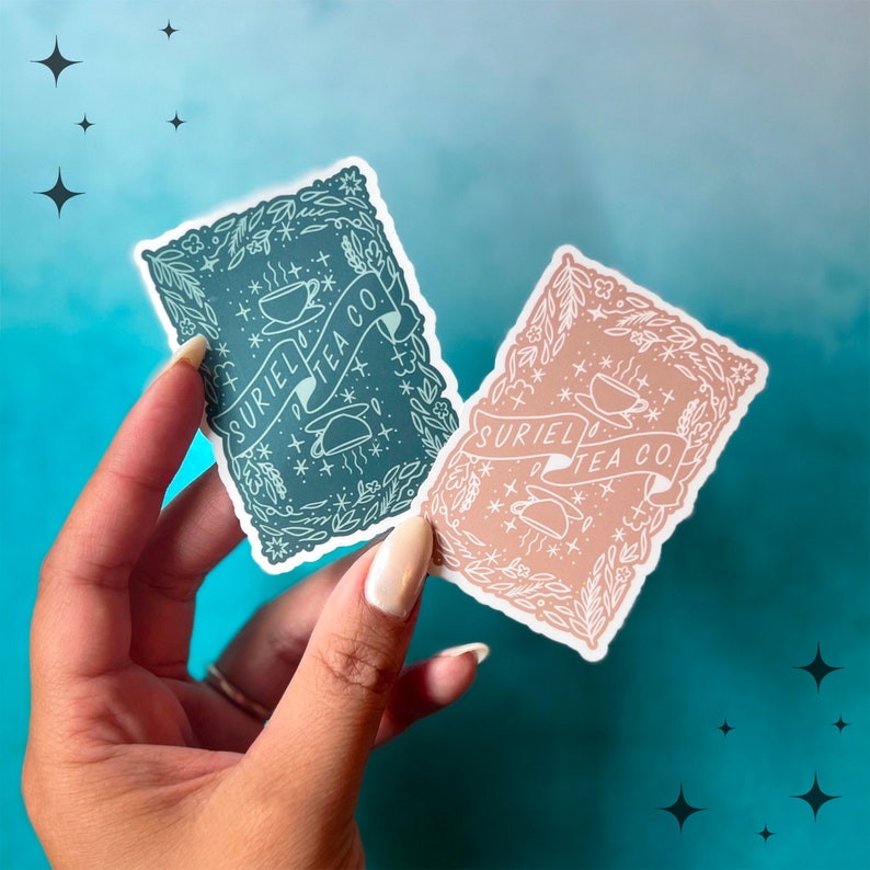 May include: Two rectangular stickers with floral borders and the text "SURIEL TEA CO." are held in a hand. One sticker is teal, the other is a muted peach colour. Both stickers feature a teacup design and star accents.