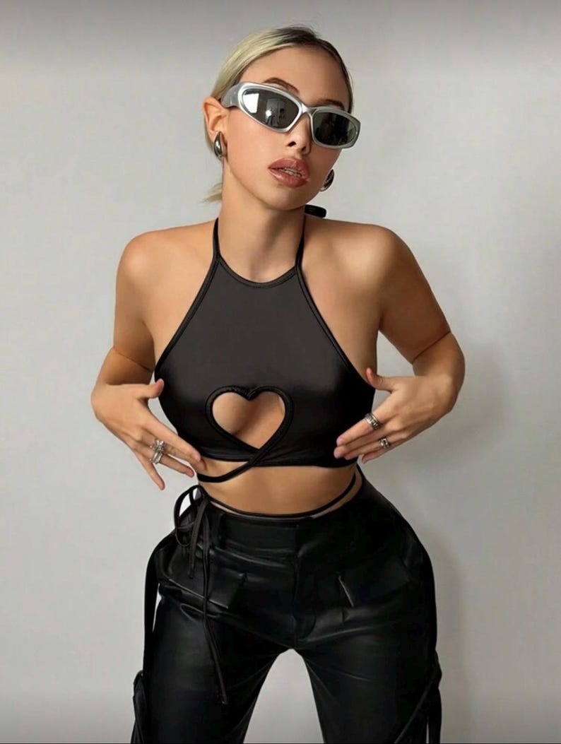 May include: A black halter top with a heart-shaped cutout and tie closure. The top is paired with black leather pants. The model is wearing silver sunglasses and rings.