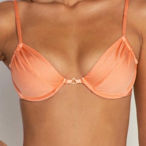 May include: Peach-colored bra with adjustable straps and a triangle-shaped gold-tone clasp. The bra has a soft, silky texture and a gathered design at the top of the cups. The bra is a lingerie item.