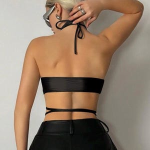 May include: A black halter top with a bandeau-style bodice and wrap-around ties. The top is paired with black leather pants. The halter top is made of a shiny material.
