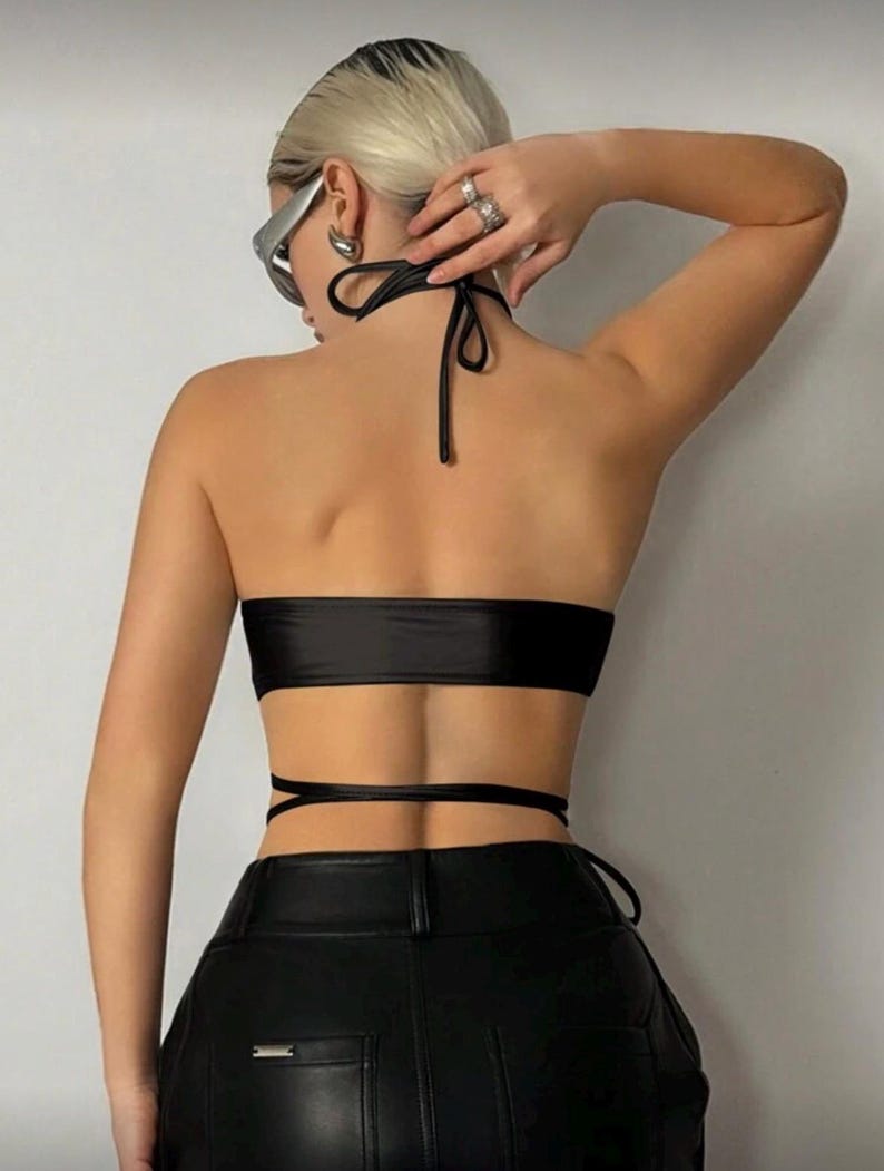 May include: A black halter top with a bandeau-style bodice and wrap-around ties. The top is paired with black leather pants. The halter top is made of a shiny material.