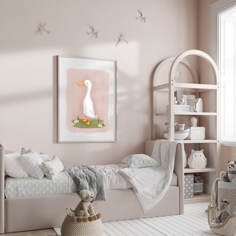 May include: A white duck with an orange beak stands on a pink and green background in a framed illustration. The illustration is hanging on a light pink wall in a nursery with a white crib and a wooden shelf with toys.