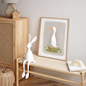 May include: A white rabbit figurine with long legs sits on a light wood bench. A framed print of a white duck with an orange beak is on the wall behind the bench. A rainbow-colored wooden toy is on the bench next to a white notebook.