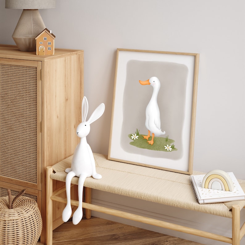 May include: A white rabbit figurine with long legs sits on a light wood bench. A framed print of a white duck with an orange beak is on the wall behind the bench. A rainbow-colored wooden toy is on the bench next to a white notebook.