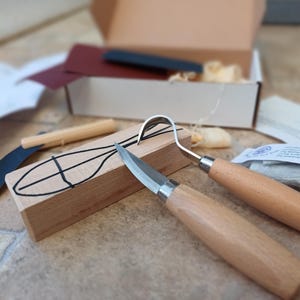 May include: A wood carving set featuring a hook knife, detail knife, and curved blade. The set includes a block of wood with a design, sandpaper, and a small box. Tools have wooden handles. The wood block has a design drawn on it.