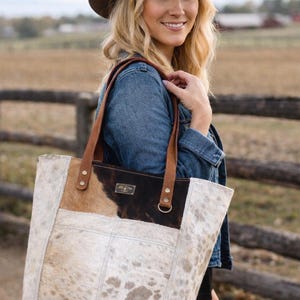 May include: A cowhide tote bag with brown leather handles and accents. The bag features a patchwork design with white, tan, and dark brown sections. The woman in the image is wearing a brown cowboy hat and a denim jacket.