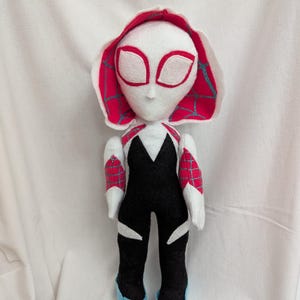 May include: A handmade plush doll of a superhero character. The doll features a white head with large eye cutouts, a red and white hood, and a black and white bodysuit. The doll's shoes are light blue.