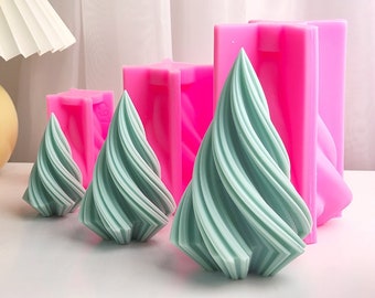 May include: Three teal green swirl-shaped candle molds in different sizes. The molds are made of pink silicone and are sitting on a white surface.