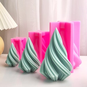 May include: Three teal green swirl-shaped candle moulds in different sizes. The moulds are made of pink silicone and are sitting on a white surface.