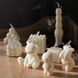 May include: An assortment of ivory-coloured candles in diverse forms, such as a bear, astronaut, honeycomb, and a figure. The candles are displayed on a wooden surface, with a tall, segmented candle at the back. Each candle is fitted with a wick.