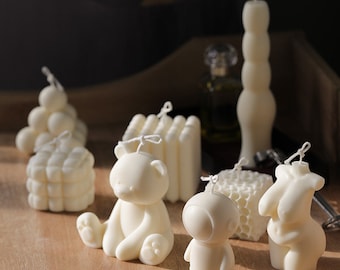 May include: A collection of ivory-colored candles in various shapes, including a bear, astronaut, honeycomb, and a figure. The candles are arranged on a wooden surface, with a tall, segmented candle in the background. Each candle has a wick.