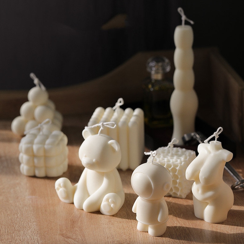 May include: An assortment of ivory-coloured candles in diverse forms, such as a bear, astronaut, honeycomb, and a figure. The candles are displayed on a wooden surface, with a tall, segmented candle at the back. Each candle is fitted with a wick.