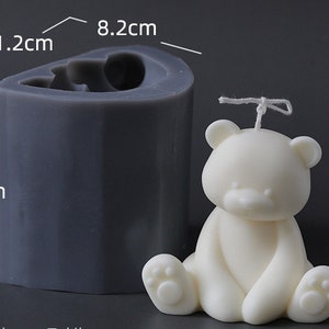 May include: A white bear-shaped candle next to a dark grey silicone mould. The candle is 10.2 cm tall. The mould is 11.2 cm wide and 8.2 cm deep. The candle has a wick and is sitting down.