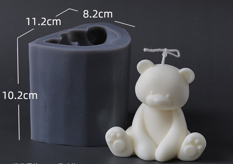 May include: A white bear-shaped candle next to a dark grey silicone mould. The candle is 10.2 cm tall. The mould is 11.2 cm wide and 8.2 cm deep. The candle has a wick and is sitting down.