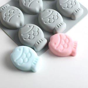 Puede incluir: Un molde de silicona con seis cavidades en forma de pez. Se muestran dos jabones en forma de pez, uno en rosa y otro en azul claro.