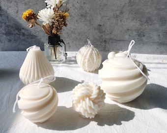May include: Five white, decorative, scented candles in various shapes, including a sphere, a pear, and a swirl design. The candles are arranged on a white surface.