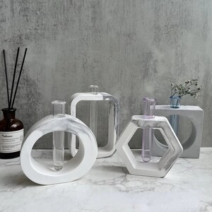 Peut inclure: Quatre vases en béton blanc de formes géométriques différentes avec des tubes en verre transparent pour les fleurs. Les vases ont un effet marbré et sont disposés sur une surface blanche.