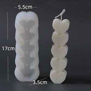 May include: A white candle shaped like stacked hearts, approximately 17 cm tall and 3.5 cm wide. Next to it is a mould, 17 cm tall and 5.5 cm wide, with heart-shaped indentations. The candle has a string wick.