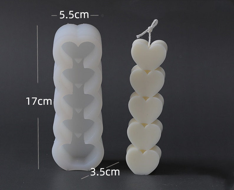 May include: A white candle shaped like stacked hearts, approximately 17 cm tall and 3.5 cm wide. Next to it is a mould, 17 cm tall and 5.5 cm wide, with heart-shaped indentations. The candle has a string wick.