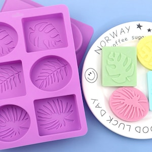 Può includere: Stampo in silicone viola con sei cavità per realizzare sapone o candele. Le cavità sono a forma di foglie e cerchi. Ci sono quattro saponette finite su un piatto bianco con il testo "Good Luck Day" e "Norway" con una grafica di tazza da caffè e zuccheriera.