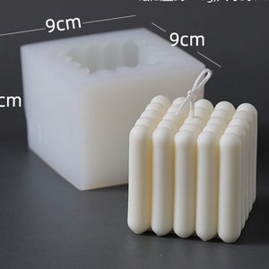 May include: A white, square candle with a ribbed design, next to a silicone mould. The candle measures approximately 9cm wide and 7.2cm tall. The mould is 9cm wide and 7.2cm tall. The candle has a wick.