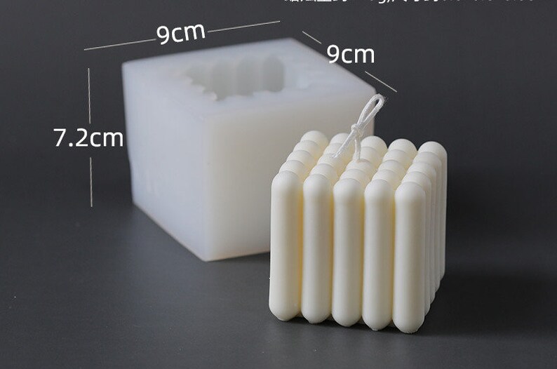 May include: A white, square candle with a ribbed design, next to a silicone mould. The candle measures approximately 9cm wide and 7.2cm tall. The mould is 9cm wide and 7.2cm tall. The candle has a wick.