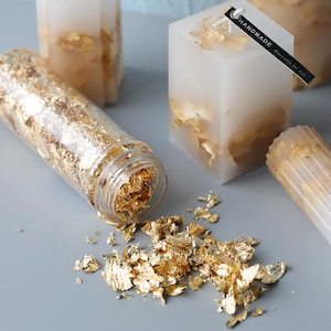 May include: A clear glass jar filled with gold leaf flakes, with some flakes scattered on a gray surface. The jar is next to a white candle with gold leaf flakes embedded in it. The candle has a black tag that says "HANDMADE especially for you".