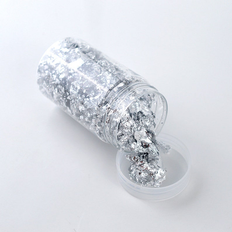 May include: A clear plastic jar filled with silver leaf flakes. The jar is open and the flakes are spilling out onto a white surface.