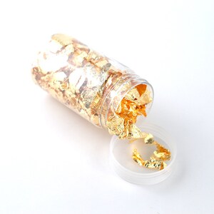 May include: A clear glass jar filled with gold leaf flakes. The jar is open and some of the flakes are spilling out.