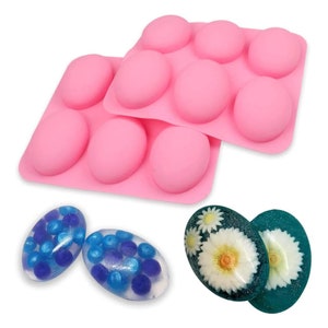 Puede incluir: Dos moldes de silicona rosa para hacer barras de jabón ovaladas. Los moldes están apilados uno encima del otro. Hay tres barras de jabón ovaladas en la imagen, una con purpurina azul, otra con purpurina verde y flores prensadas, y otra con purpurina azul y morada.