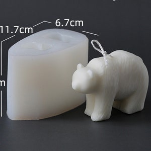 May include: A white bear-shaped candle and its silicone mould. The candle has a wick and is 6.7 cm wide, 7.7 cm tall, and 11.7 cm long. The mould is the same shape and size as the candle. The background is a dark grey.